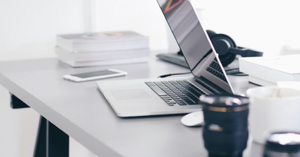 Half-closed laptop on a table with a smartphone, books, headphones and camera lenses.